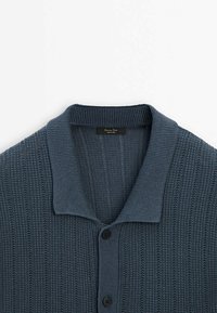Cardigan bleu en tricot avec col en côtes et fermeture à boutons, présentant des motifs verticaux texturés et une étiquette de marque à l'intérieur du col.