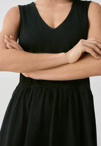 Schwarzes ärmelloses Kleid mit V-Ausschnitt, das ein figurbetontes Oberteil und einen ausgestellten Rock hat. Hergestellt aus weichem Stoff mit glatter Textur.
