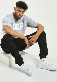 Halli lühikeste varrukatega hall t-särk, millel on valge "DEF" tekst, mustad kitsenevad püksid ja valged Nike Air Force 1 tossud, istub valgel kuubikul.