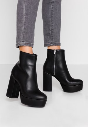 Bottines en cuir noir avec un talon épais et haut, une semelle compensée et des détails de fermeture éclair sur le côté, assorties à un jean en denim gris.