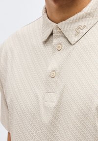 Close-up van een beige poloshirt met patroon, drie knopen en een klein reliëflogo op de kraag, gedragen door een persoon.