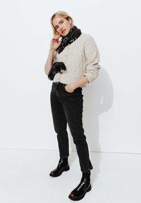 Pull en maille beige avec boutons, jean noir taille haute et bottines noires avec accents marron, accessoirisé d'une écharpe noire.