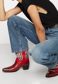 Rote Leder-Ankle-Boots mit schwarzen Flammenmustern, spitzer Zehenform und niedrigem Blockabsatz. Kombiniert mit hellblauen, cropped Jeans.
