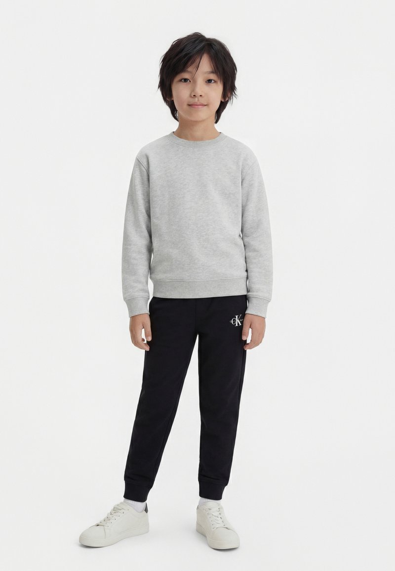 Παιδί όρθιο, φοράει ανοιχτό γκρι φούτερ, μαύρο παντελόνι με λογότυπο στον αριστερό μηρό, λευκές κάλτσες και λευκά αθλητικά παπούτσια σε λευκό φόντο.