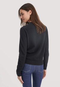 Schwarzer Strickpullover mit rundem Ausschnitt und langen Ärmeln. Rippbündchen an Saum und Ärmeln, glatte Textur, taillierte Passform. Getragen mit blauen Jeans.