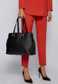 Sac cabas en cuir noir à finition texturée, avec des accents de quincaillerie dorée et deux poignées robustes. Porté par une personne en costume rouge et talons noirs.