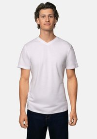 Ung mand med kort, bølget hår iført en ensfarvet hvid V-udskæring T-shirt og mørke jeans, stående foran en lys ensfarvet baggrund.
