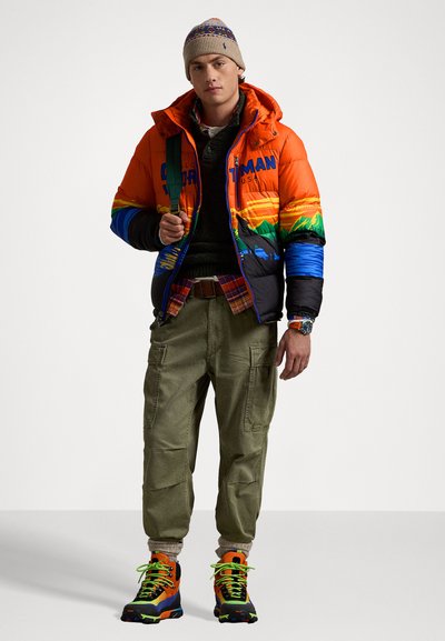 Männer mehrfarbige Pufferjacke mit Berggrafiken, kombiniert mit olivfarbenen Cargo-Hosen und lebhaften Turnschuhen mit Neon-Schnürsenkeln.