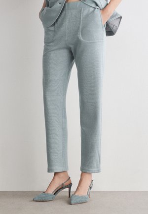 Femme portant un pantalon gris clair fuselé avec poches avant et des talons slingback tissés assortis, debout, les mains dans les poches.