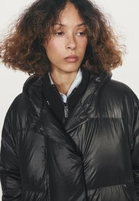 Černý péřový kabát s lesklým, texturovaným povrchem, kapucí, diagonálním zipem a nadměrným střihem s měkkou vnitřní podšívkou.