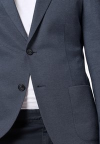 Granatowy żakiet o teksturowanej fakturze z dwoma czarnymi guzikami, jednym kieszonką i dopasowanym krojem. Pod spodem widoczna jest biała koszula.