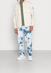 Light beige button-up shirt with two chest pockets layered over a green t-shirt, paired with blue and white tie-dye jeans and white sneakers.
