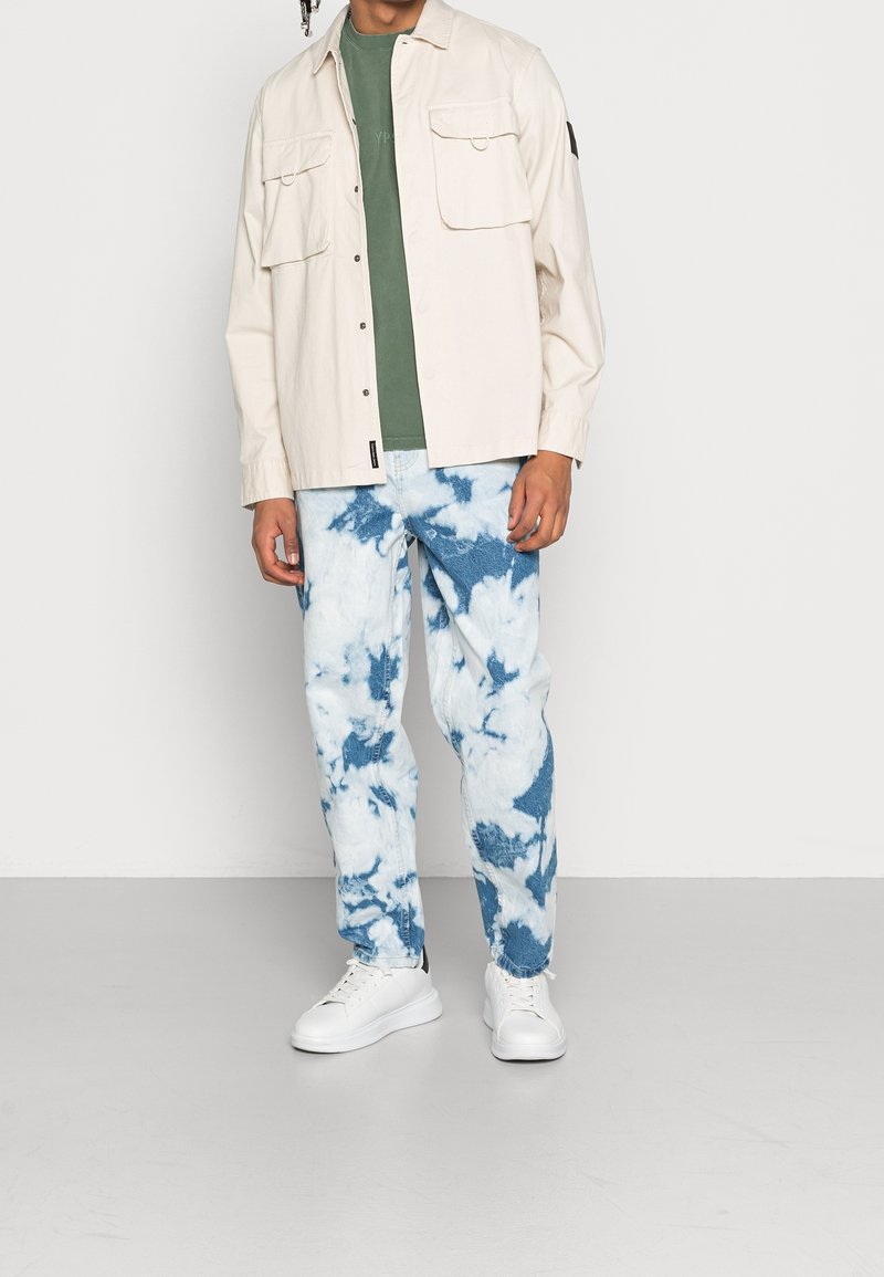 Light beige button-up shirt with two chest pockets layered over a green t-shirt, paired with blue and white tie-dye jeans and white sneakers.