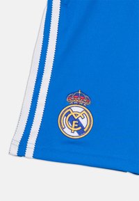 Pantalones cortos deportivos azules con franjas laterales blancas y un escudo bordado del Real Madrid con una corona dorada en la parte inferior izquierda.