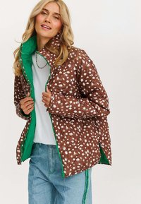 Brązowa kurtka puchowa w białe kropki, z zielonym wykończeniem, zamkiem błyskawicznym i bocznymi rozcięciami, noszona na białej bluzce i zestawiona z niebieskimi dżinsami.