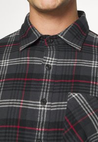 Schwarz-grau kariertes Flanellhemd mit roten Akzenten, Knopfleiste vorne, klassischem Kragen und Brusttasche. Die Baumwollstruktur ist sichtbar.