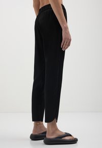 Personne portant un pantalon noir à taille élastique avec des fentes sur les côtés, des sandales noires rembourrées et un bracelet de cheville en chaîne dorée sur un fond blanc.