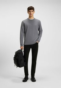 Camisa gris de manga larga con cuello redondo, pantalones negros y mochila negra con acentos texturizados, de pie sobre un fondo neutro.