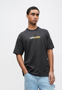 Μαύρο T-shirt με κοντά μανίκια και στρογγυλή λαιμόκοψη, διακοσμημένο με ένα γραφικό λογότυπο σε λευκό και κίτρινο. Συνδυάζεται με ανοιχτόχρωμα σορτς από τζιν.