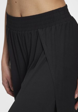 Nahaufnahme einer Person, die schwarze, lockere Hosen mit elastischem Bund und überlappendem Stoffdetail in Hüfthöhe trägt.