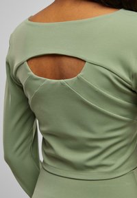 Hosszú ujjú felső tompított zöld anyagból, kivágott hátpánttal és a varrások mentén ráncolt részletekkel. Puha textúra és elegáns dizájn.