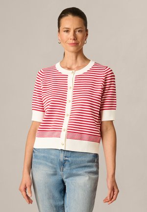 Woman with brown hair in a low ponytail wearing gold hoop earrings, a red and white striped short-sleeve buttoned cardigan, and light blue jeans.