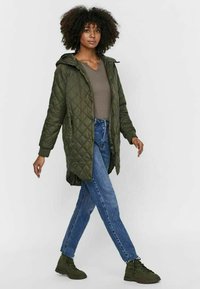 Olivgrön vadderad jacka med huva, två sidofickor, långa ärmar och knä-lång design, i kombination med blå jeans och gröna stövlar.
