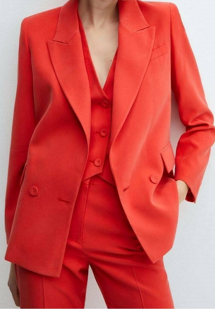 Femme portant un costume tailleur rouge vif avec un blazer croisé, un gilet assorti et un pantalon taille haute, main dans la poche.
