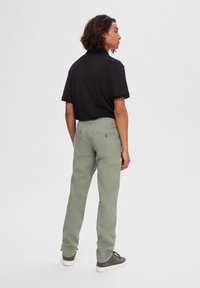 Camiseta polo negra de manga corta, pantalones de algodón verde claro, vista trasera. Los pantalones presentan dos bolsillos traseros con detalles de botones, combinados con zapatos grises.