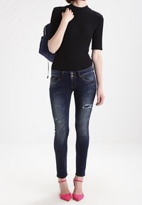 Top negro de manga corta, ajustado; jeans ajustados azul oscuro con detalles desgastados; zapatos de tacón alto rosas; mochila azul en el hombro izquierdo.