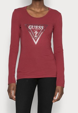 Vrouw draagt een getailleerd langemouws shirt in bordeauxrood met een zilveren pailletten "GUESS U.S.A" driehoekig logo op de borst.