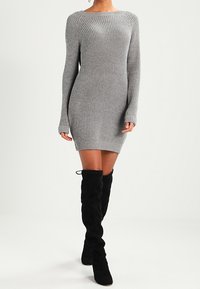 Kvinna klädd i en figurnära grå ribbad stickad klänning med långa ärmar och svarta mocka stövlar över knäna, som går framåt.