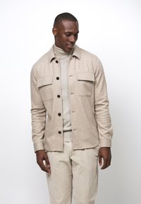 Beige knoopjacket met twee borstzakken en een rechte pasvorm, gedragen over een lichte coltrui, gecombineerd met crèmekleurige corduroy broek.