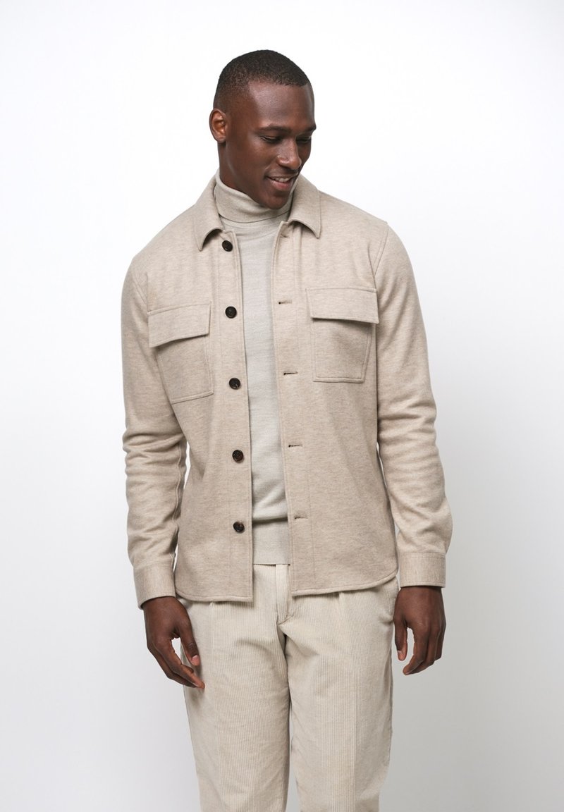 Beige knoopjacket met twee borstzakken en een rechte pasvorm, gedragen over een lichte coltrui, gecombineerd met crèmekleurige corduroy broek.