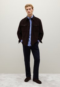 Bruine corduroy jas met knoopzakken, laag over een blauw-wit gestreept shirt. Gepaard met donkere broeken en bruine schoenen.