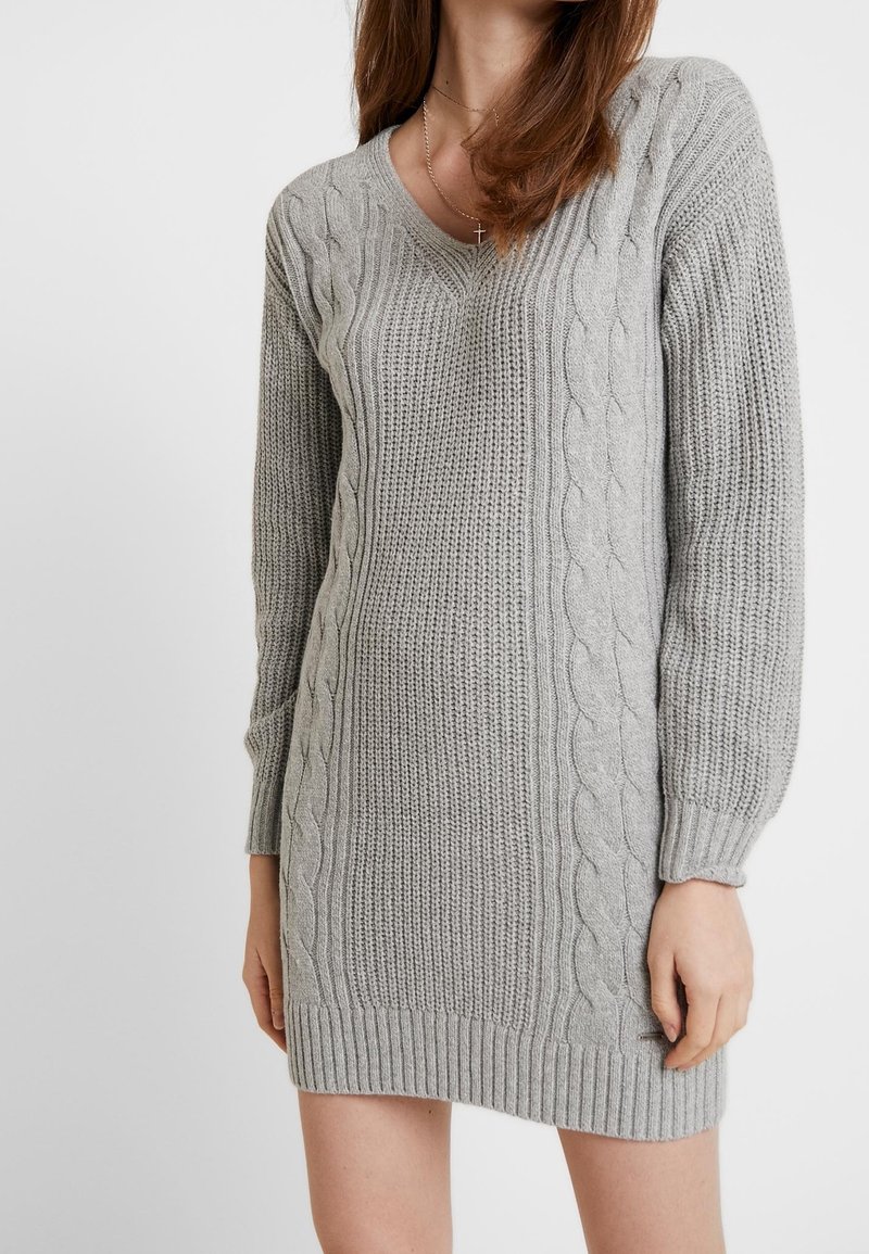 Grå stickad klänningströja med V-ringning, med kabelmönster längs sidorna, ribbad nederkant och långa ärmar. Mjuk textur.