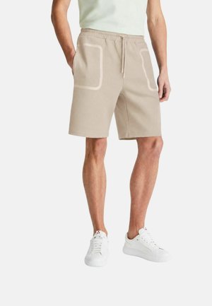 Uomo con pantaloncini beige fino al ginocchio con tasche bordate di bianco e scarpe da ginnastica bianche, in piedi con le mani rilassate lungo i fianchi.