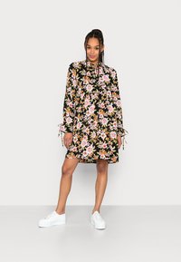 Vestido floral com fundo preto, apresentando flores em tons de rosa e laranja, mangas longas com cordões e um corte solto. Combinado com ténis brancos.