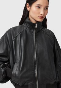 Černá kožená bomber bunda s vysokým límcem, kovovým zipem a bočními kapsami. Má žebrované manžety a hladkou, lesklou texturu.