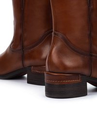 Une paire de bottes en cuir marron avec des talons en bois sombre empilés et des coutures détaillées sur un fond blanc.