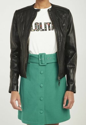 Femme portant une veste en cuir noire sur un t-shirt blanc à motifs floraux avec l'inscription « LOLITA » et une jupe verte taille haute avec des boutons et une ceinture assortie.