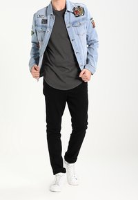 Světle modrá džínová bunda s různými nášivkami, šedé tričko pod ní, černé kalhoty a bílé tenisky. Neformální oblečení s výraznými detaily.