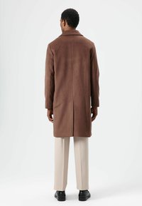 Bruine, lange jas met een gladde textuur en klassieke kraag, voorzien van een rechte snit en een centrale achtersplit, gecombineerd met beige broek.