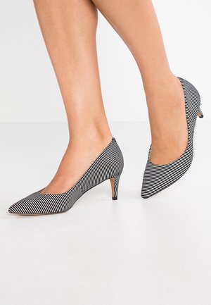 Fødder iført sorte og hvide stribede kitten heel-sko med spids tå, stående på et hvidt gulv med en ensfarvet baggrund.