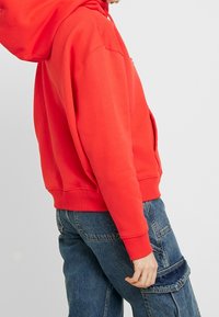Red cotton hoodie with a kangaroo pocket and ribbed cuffs. Worn with blue denim cargo pants featuring side pockets and a distressed texture.