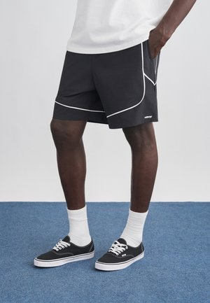 Άτομο που φοράει μαύρο σορτς με λευκή επένδυση, λευκές κάλτσες και μαύρα χαμηλά αθλητικά παπούτσια που στέκεται σε μπλε χαλί.