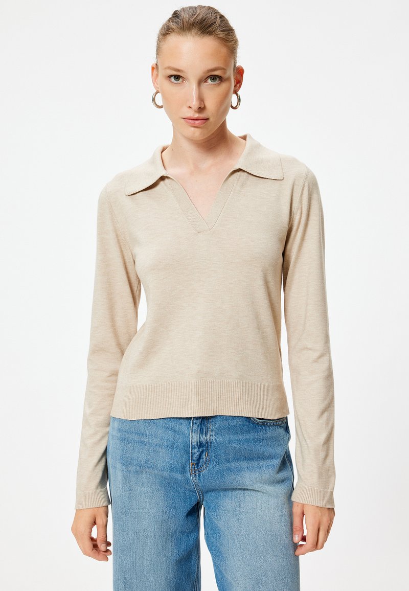 Beżowy pulower z długim rękawem, z dekoltem w kształcie litery V i kołnierzykiem, wykonany z miękkiej tkaniny. Posiada ściągacze przy hemie i mankietach, zestawiony z jasnoniebieskimi dżinsami.