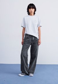 Lys grå t-skjorte med logo, kombinert med løstsittende mørk grå jeans. Modellen har på seg svart-hvite joggesko mot en blå bakgrunn.