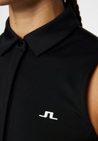 Svart polo-tröja i ett slätt tyg, med en knappat krage och en vit logotyp på bröstet, med en elegant, figurnära silhuett.