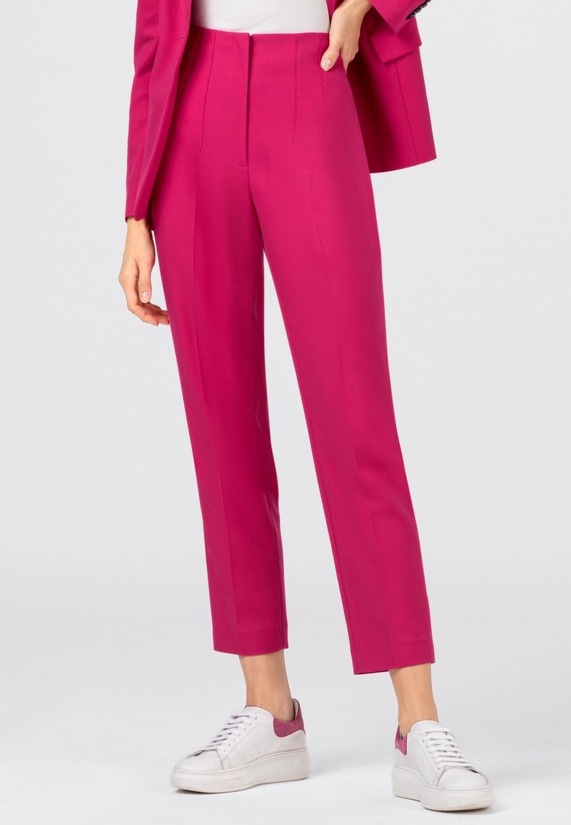 Femme portant un pantalon rose vif tailleur longueur cheville avec un blazer assorti et des baskets blanches avec des touches roses.